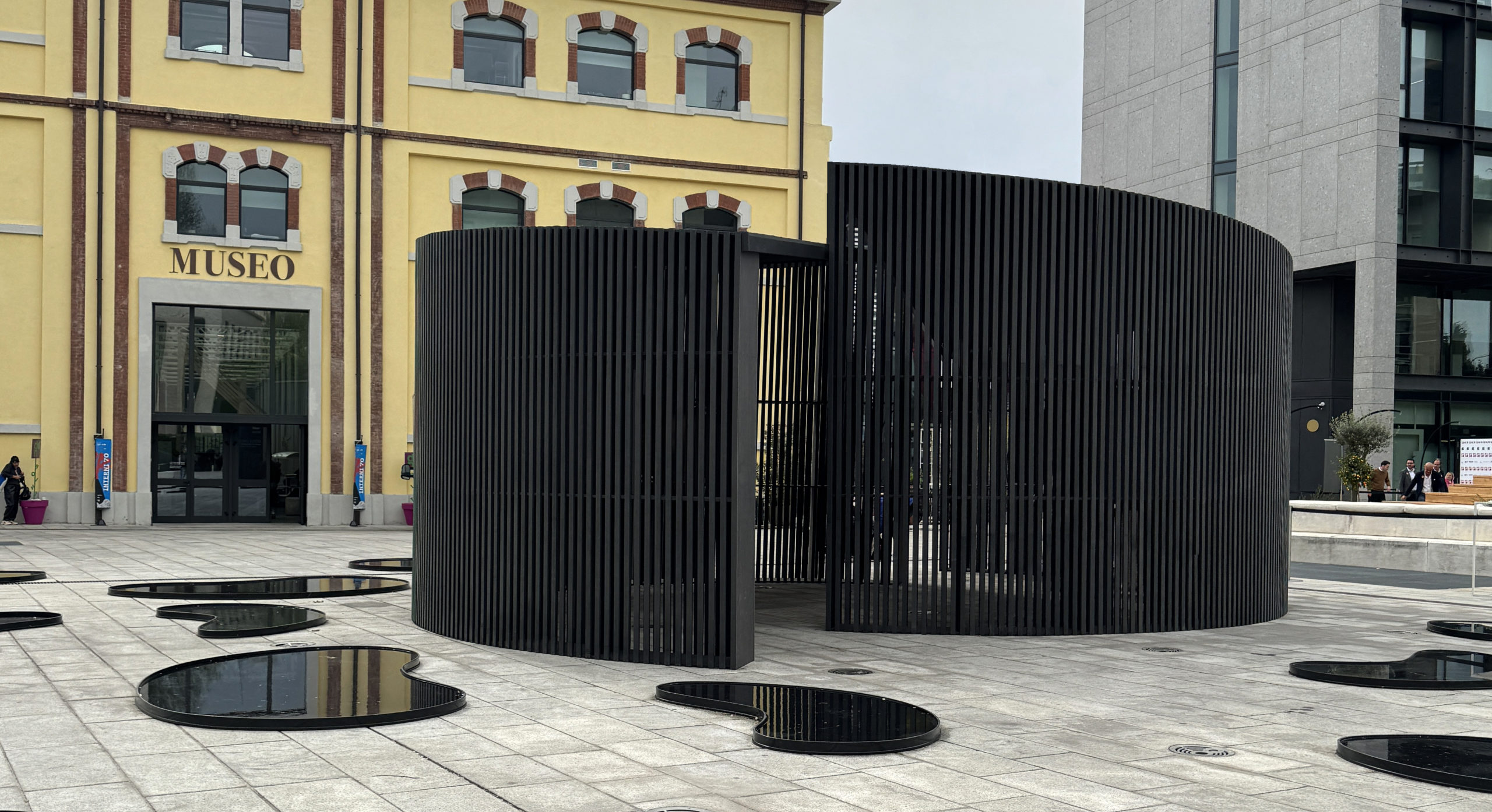 Black slatted AD AQUAM pavilion outside the ADI Design Museum, with reflective water-shaped elements on the ground.